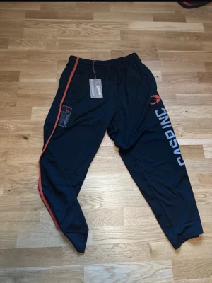 Svarta träningsbyxor från GASP - Säljer ett par svarta träningsbyxor från GASP med orange sidostripes och logga på benet. Byxorna har resår i midjan med dragsko och är tillverkade i ett lätt och bekvämt material, perfekt för gymmet eller sportaktiviteter. De har inte prislappen kvar utan de där är gamla bilder men har använt de en gång så de ser lika dana ut som på bilden