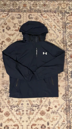 Marinblå vindjacka från Under Armour - Snygg marinblå vindjacka från Under Armour med huva och dragkedja framtill. Jackan har elastiska muddar vid ärmsluten, två sidofickor med dragkedja och reflekterande detaljer. Perfekt för blåsiga dagar och sportiga outfits.