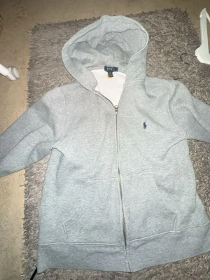 Grå hoodie från Stone Island - Säljer en grå hoodie från Stone Island med dragkedja framtill och klassisk huva. Hoodien har den ikoniska Stone Island-loggan på ärmen och praktiska fickor framtill. Perfekt för dig som gillar streetwear och vill ha en stilren look.