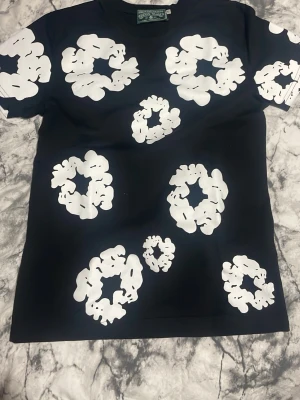 Svart t-shirt från Denim Tears - Säljer en svart t-shirt från Denim Tears med stora vita blommiga tryck över hela tröjan. T-shirten har rund hals och korta ärmar. Materialet är mjuk bomull som känns skön mot huden. Perfekt för dig som vill sticka ut med en unik och trendig look.