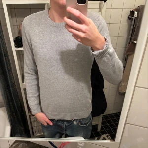 Ljusgrå stickad tröja med rund hals - En stilren ljusgrå stickad tröja med rund halsringning och ribbade muddar vid ärmslut och nederkant. Tröjan har en liten diskret etikett nertill och passar perfekt till jeans för en avslappnad look.