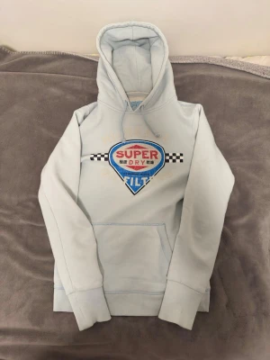 Ljusblå Superdry hoodie med tryck - Ljusblå hoodie från Superdry med stort broderat och tryckt motiv på bröstet i blått, rött, svart och vitt. Klassisk huva med dragsko och magficka framtill. Mjuk bomullsblandning och ribbade muddar vid ärmar och nederkant. Perfekt för en avslappnad streetwear-look.