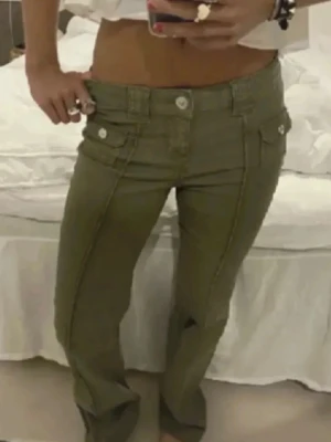 Olivgröna lågmidjade bootcut jeans - Säljer ett par olivgröna jeans med låg midja och bootcut passform. Jeansen har dekorativa fickor framtill med knappar och markerade sömmar längs benen. Perfekta för dig som gillar en retro och trendig look. Lite olika bilder med olika ljus!