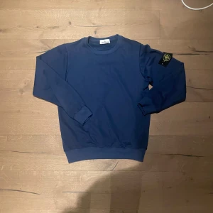 Blå Stone Island sweatshirt - Snygg marinblå sweatshirt från Stone Island med klassisk rund hals och ikonisk patch på vänster ärm. Tröjan är tillverkad i mjuk bomull och har ribbade muddar vid ärmslut och nederkant. Perfekt för dig som gillar streetwear och vill ha något stilrent.