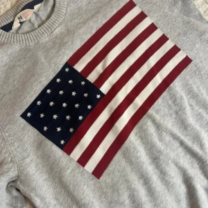 Grå stickad tröja med USA-flagga - Snygg grå stickad tröja från H&Mmed stor amerikansk flagga på bröstet. Tröjan har rund halsringning och långa ärmar!