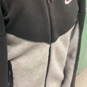 Nike tech  - Nike tech i tvåfärgad design med svart upptill och ljusgrå nedtill. Tröjan  har dragkedjor vid fickorna och en sportig look med synliga sömmar. Perfekta för chill eller träning, med klassisk Nike-logga på höften.