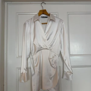 Vit klänning i satin från Zara - Supersnygg vit kortklänning från Zara i glansig satin med omlott-design och krage. Klänningen har långa ärmar med breda manschetter och en figurnära passform som ger en elegant siluett. Perfekt för dig som gillar stilrena och trendiga plagg.