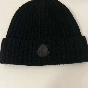 Svart ribbstickad mössa från Moncler - Svart ribbstickad mössa från Moncler med uppvikt kant och diskret Moncler-logga framtill. Klassisk form och stilren design som passar perfekt till kyliga dagar. Tillverkad i mjukt och värmande material för extra komfort.