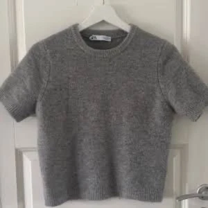 Grå stickad kortärmad Zara tröja - Säljer en grå stickad tröja från Gina Tricot med korta ärmar och rund hals. Tröjan har en ribbad kant vid hals, ärmslut och nederkant. Perfekt för dig som gillar enkel och stilren look.