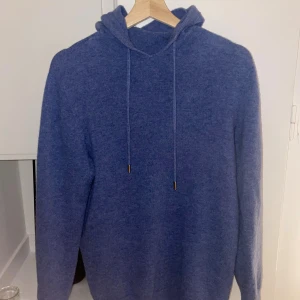 Blå ull-hoodie - Blå hoodie i mjukt stickat material med huva och snörning. Tröjan har långa ärmar och ribbade muddar vid ärmslut och nederkant. Perfekt för kyliga dagar och har en chill, enkel look. Passar bra mellan 173-184. Har några små defekter men inte tillräckligt för att se på.