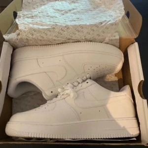 Nike Air Force 1 helvita sneakers - Nike Air Force 1 sneakers i helvit färg med klassisk låg siluett. Skorna har snörning, perforerad tå för ventilation och en platt, robust sula med cirkulärt mönster. Tillverkade i skinn med mjuk vadderad insida för extra komfort. Perfekta för dig som gillar clean och stilren look.