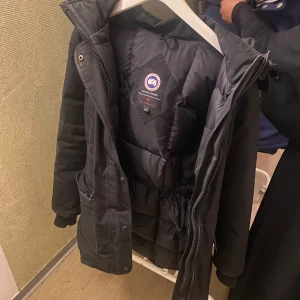Svart dunjacka från Canada Goose M - Säljer en svart dunjacka från Canada Goose med klassisk logga på ärmen. Jackan har huva, dragkedja och knappar framtill samt elastisk midja för snygg passform. Perfekt för kalla dagar och riktigt varm. Fodrad insida och flera praktiska fickor.