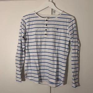 Randig långärmad tröja från Lager 157 - Vit långärmad tröja med blåa ränder från Lager 157. Tröjan har rund halsringning, knappar framtill och ett litet broderat emblem på bröstet.