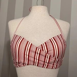 Rödvit randig bikini med knyt - Snygg bikini med rödvita ränder och omlott-topp. Toppen har smala axelband som knyts i nacken och en bred knytning i ryggen. Bikinitrosan har samma randiga mönster och klassisk passform. Endast använd en gång. Storlek M på både topp och trosa. 