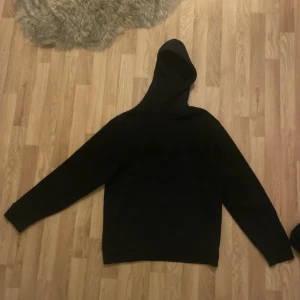 Svart hoodie med huva - En klassisk svart hoodie med huva och långa ärmar. Perfekt för att slänga på sig när det är lite kyligare ute. Enkel och stilren design utan tryck eller detaljer.