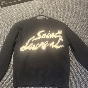 stickad Saint Laurent tröja - Svart stickad tröja från Saint Laurent med fluffigt vitt logotyptryck på bröstet. Rund hals och långa ärmar med ribbade muddar. Perfekt för dig som vill ha en cool statement piece med lyxig vibe och unik design.