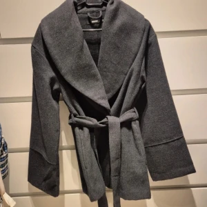 Grå omlott kappa från Bikbok - Snygg grå kappa från Bikbok i omlottmodell med bred krage och bälte i midjan. Jackan har lång ärm och en loose passform som ger en chill vibe. Perfekt för dig som gillar stilrena och enkla plagg med lite edge.