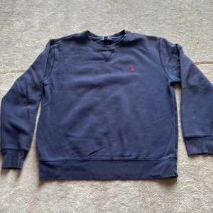 Mörkblå sweatshirt från Polo Ralph Lauren - Snygg mörkblå sweatshirt från Polo Ralph Lauren med klassisk crewneck och röd broderad logga på bröstet. Tröjan har långa ärmar, ribbade muddar och är gjord i mjuk bomull. Har ett litet hål uppe vid vänstra axeln. Se bild. 