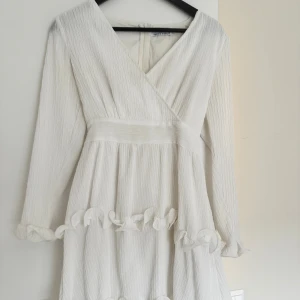 Vit omlottklänning från Dennis Maglic - Superfin vit omlottklänning från Dennis Maglic med långa ärmar, v-ringning och volangdetaljer vid ärmslut och kjol. Knytband i midjan och lätt plisserat tyg ger en romantisk vibe. Perfekt för student eller sommarens alla tillfällen! Kan knytas såväl i ryggen som fram!