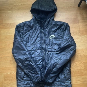 Svart quiltad pufferjacka från Nike - Svart pufferjacka från Nike med quiltad design och huva. Jackan har dragkedja framtill och ett broderat Nike-logga på bröstet. Yttermaterialet är glansigt och foder i fleece för extra värme. Perfekt för kyliga dagar.