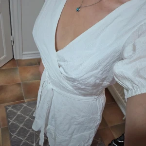 Nelly Vit Omlottklänning (Flounce Structure Dress) - Strl 36 - Säljer en fin vit omlottklänning från Nelly i storlek 36. Klänningen har en smickrande V-ringning, puffärmar och knyts i midjan för att framhäva figuren. Tyget har en lätt struktur (enligt etikett: "Flounce Structure Dress"). Den är perfekt för sommarfester, stranden, eller som en enkel och söt vardagsklänning under varma dagar. Klänningen är fodrad (100% Polyester foder) vilket minskar transparensen.