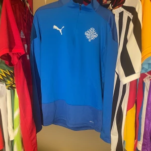 Blå Island landslags Puma half zip hoodie S Fotbollströja - Blå hoodie från Puma med half zip och lång ärm. Tröjan har vit Puma-logga och Islands märke på bröstet. Fotbollströja