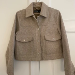 Beige kort jacka från Zara  - Stilren beige jacka från Zara med stora bröstfickor och silvriga tryckknappar. Jackan har rak passform, klassisk krage. Enkel att styla till olika outfits.