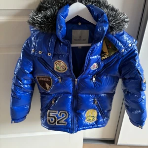 Blå Moncler dunjacka med patches - Snygg blå dunjacka från Moncler med glansig finish, coola broderade patches och fuskpäls på huvan. Jackan har dragkedjor framtill och på fickorna, samt flera detaljer som ger en sportig vibe. Perfekt för dig som vill sticka ut i vinterkylan. Storlek ca 130, dvs barn. Vet ej om den är äkta , hon jag köpte den av sa det. Men den var för liten för min son som är 10. 