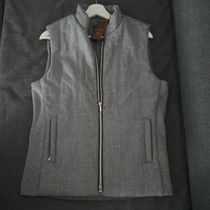Grå väst från Stenströms med dragkedja - Snygg grå väst från Stenströms med rak passform och hög krage. Västen har två fickor fram och stängs med en silverfärgad dragkedja. Materialet är mjukt och har en diskret textur, perfekt för lager-på-lager. 