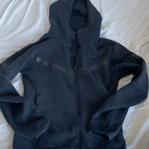 Svart Nike hoodie med dragkedja XS - Svart hoodie från Nike i storlek XS med hel dragkedja framtill och huva. Tröjan har diskret Nike-logga på bröstet och praktiska fickor med dragkedja. Perfekt för dig som gillar sportig och clean stil.
