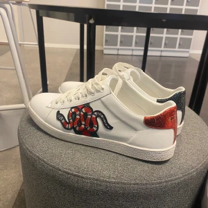 Gucci Ace sneakers med orm - Snygga vita Gucci Ace sneakers i skinn med broderad röd och svart orm på sidan. Baktill har ena skon röd och andra grön metallic-detalj med ormskinnsmönster och Gucci-logga. Klassisk låg modell med snörning och platt sula. Perfekt statement-piece för din outfit.
