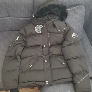 Svart pufferjacka Moose Knuckles XL - Säljer en svart pufferjacka från Moose Knuckles i storlek XL. Jackan har en fet pälskantad huva, ikoniska metall-loggan på ärmen och flera fickor med både dragkedja och knappar. Perfekt för kalla dagar och riktigt snygg streetstil.