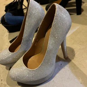 Glittriga silverpumps med hög klack - Säljer ett par riktigt snygga pumps från Lucky i storlek 35. Skorna är täckta av glittrigt silvertyg och har en hög klack samt plattform framtill. Perfekta för dig som vill sticka ut med extra bling. Rund tå och klassisk pumpsform.