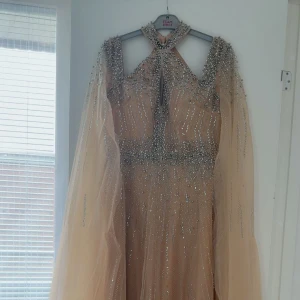 Champagnefärgad glittrig långklänning - Magisk långklänning i champagnefärg med glittriga paljettdetaljer och transparenta ärmar. Klänningen har en halterneck-ringning med snygg cutout framtill och ett svepande, voluminöst kjolparti. Perfekt för dig som vill sticka ut på bal eller fest.