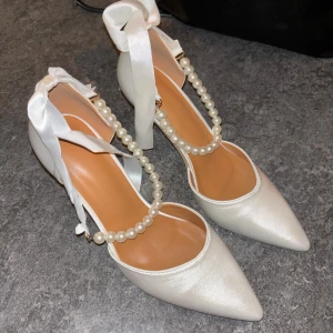 Vita pumps med pärlband och rosett - Eleganta vita pumps med spetsig tå, klack och pärlband över foten. Skorna har en fin rosett i sidenband som knyts runt ankeln och är tillverkade i ett glansigt satinliknande material. Perfekta för dig som vill sticka ut med en chic och feminin look.