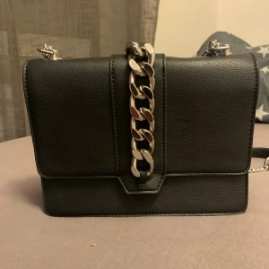 Svart axelväska med kedjedetalj - Stilren svart axelväska i skinnimitation med grov silverfärgad kedja framtill. Väskan har en fyrkantig form och justerbar axelrem i kedja. Perfekt för dig som vill ha en snygg och trendig accessoar till din outfit.