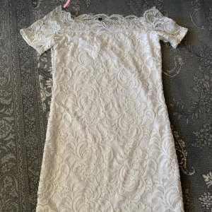 Vit spetsklänning med offshoulder - Superfin vit kortklänning i spets med offshoulder och korta ärmar. Klänningen har ett romantiskt mönster och är perfekt för sommarens alla tillfällen. Snygg passform och vackra spetsdetaljer längs hela klänningen.
