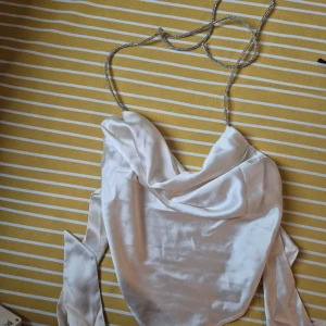 Vit satintopp med glittriga band - Säljer en vit satintopp med glittriga smala axelband och draperad halsringning. Toppen har en croppad passform och knytband i ryggen för extra detalj. Perfekt för dig som gillar att sticka ut med stilrena detaljer.