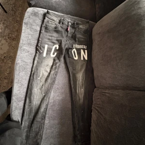Svarta Dsquared2 ICON jeans, strl 44 - Snygga svarta jeans från Dsquared2 med stor vit 'ICON'-print framtill och coola slitningar samt färgstänk. Bakficka med dragkedjedetalj och Dsquared2-lapp i rött och svart. Slim fit-modell i stretchig bomull för en modern look.