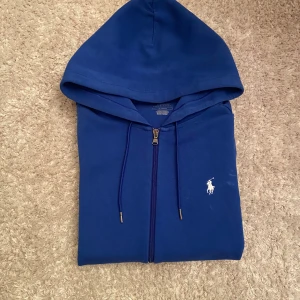Blå hoodie från Polo Ralph Lauren - Snygg blå hoodie från Polo Ralph Lauren med klassisk vit broderad logga på bröstet. Tröjan har huva med snörning och dragkedja framtill. Perfekt för dig som gillar stilrena och sportiga plagg. Materialet är mjuk bomull som känns skönt mot huden. Loggan på bröstet kanske ser lite gul ut men det är bara på grund av ljuset i bilden, i verkligheten är den vit. 