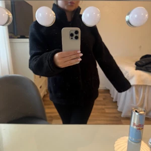 Svart teddyjacka med dragkedja - Mysig svart teddyjacka med hög krage och hel dragkedja framtill. Jackan har två sidofickor och en extra ficka med dragkedja på bröstet. Perfekt för kyliga dagar och har en loose passform som ger en chill vibe.