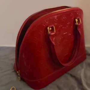 Röd handväska från Louis Vuitton - Snygg röd handväska från Louis Vuitton i glansigt skinn med klassiskt LV-mönster. Väskan har guldfärgade detaljer, dubbla handtag och dragkedja. Medföljer matchande hänglås och nyckel i guld. Perfekt storlek för dina essentials och ger en lyxig vibe. 