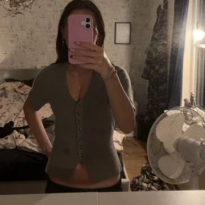 Grå kortärmad topp med knappar - Grå topp med v-ringning och korta ärmar. Toppen har en rad knappar framtill och är lätt croppad. Enkel och stilren design som passar till mycket. Mjuk och bekväm, perfekt för en avslappnad look.