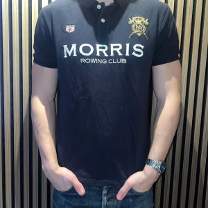 Svart piké från Morris Rowing Club - Svart pikétröja från Morris Rowing Club med broderade detaljer och emblem på bröstet. Klassisk krage, korta ärmar och knappar framtill. Stor logotyp och text på framsidan. Skön och stilren, perfekt för en sportig look. Storlek L men passar M.