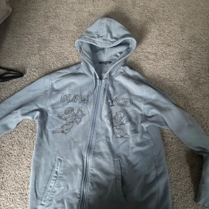Ljusblå hoodie från Blinglucid - Ljusblå hoodie från Blinglucid med dragkedja, huva och två fickor. Framsidan har glittriga motiv av änglar och texten 'BLING LUCID' i stenar.
