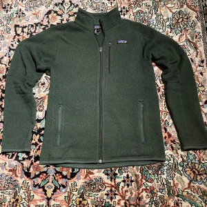 Mörkgrön fleecejacka från Patagonia - Mörkgrön fleecejacka från Patagonia i storlek S. Jackan har hel dragkedja framtill, två sidofickor med dragkedja och en bröstficka. Tillverkad i 100% återvunnen polyester