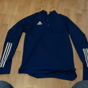 Blå Adidas långärmad träningströja - Blå långärmad träningströja från Adidas med klassiska vita ränder på ärmarna och Adidas-logga på bröstet. Tröjan har half zip framtill och är tillverkad i polyester, perfekt för sport eller chill. Snygg och sportig look!