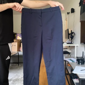 Mörkblå kostymbyxor med raka ben - Snygga mörkblå kostymbyxor med raka ben och klassiska fickor bak. Byxorna har hällor för bälte och stängs med dragkedja och knapp. Perfekta för en clean och stilren look. Materialet känns mjukt och har en lätt struktur.