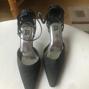 Svarta pumps med nitar och spetsig tå - Snygga svarta pumps från Juse 4U med spetsig tå och smal klack. Skorna har coola nitar och remmar runt hälen samt justerbart spänne vid ankeln. Perfekta för dig som vill sticka ut med en edgy stil.