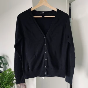 Svart kofta från H&M - En klassisk svart kofta från H&M med v-ringning och knäppning framtill. Långärmad modell med ribbade muddar vid ärmslut och nederkant. Perfekt att slänga över en t-shirt eller blus. Storlek M. 83 % ekologisk bomull, 17 % polyamid. Axelbredd: 36 cm | Längd: 55 cm | Ärmlängd: 56 cm. I gott skick. Kan hämtas i Kista, Stockholm, eller skickas.